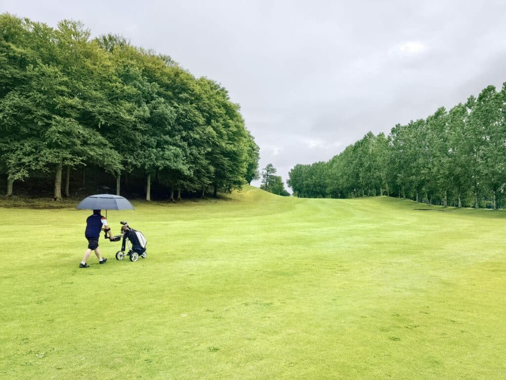 Man with umbrella and golf cart on a lush green golf course in Tuve, Sweden representing Best Golf Umbrella. 