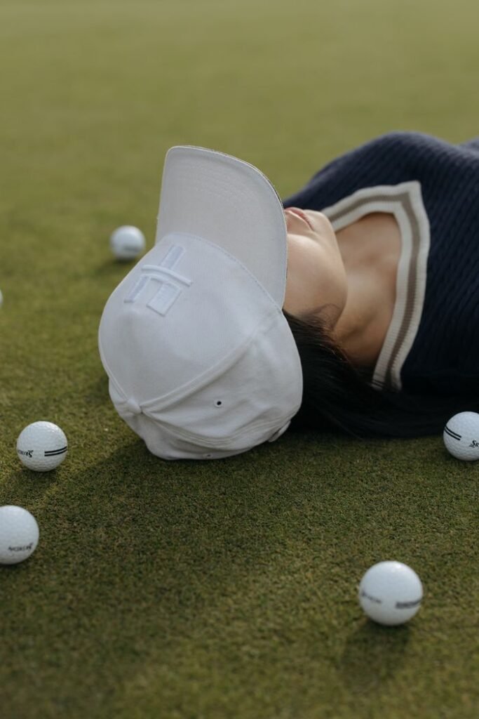 pexels-photo-6230532-6230532 A woman in a cap lying on a golf course surrounded by scattered golf balls.